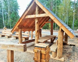 Drewniana wiata dębowa z dachem dwuspadowym i ławostołem z litego drewna w stylu rustykalnym, ustawiona na terenie leśnym jako element małej architektury parkowej. W tle widoczne ławki i kosze drewniane – kompleksowe wyposażenie strefy odpoczynku w otoczeniu natury.