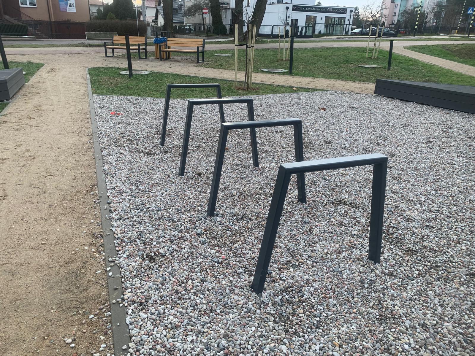 STOJAK NA ROWER (4) Stalowy stojak rowerowy typu odwrócone U na placu miejskim, zamontowany w podłożu żwirowym wśród małej architektury miejskiej