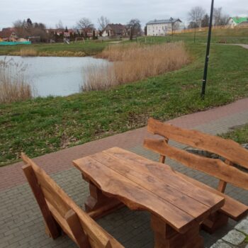 lawostol prosty_Kwidzyn Balaton (5) Zestaw ławostołów z naturalnego drewna ustawiony na brukowanej ścieżce przy zbiorniku wodnym. Drewniane ławki i stół o masywnym i rustykalnym charakterze, wkomponowane w otoczenie zieleni i krajobrazu miejskiego z alejkami i budynkami w tle