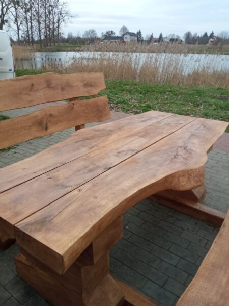 lawostol prosty_Kwidzyn Balaton (8) Zestaw ławostołów z naturalnego drewna ustawiony na brukowanej ścieżce przy zbiorniku wodnym. Drewniane ławki i stół o masywnym i rustykalnym charakterze, wkomponowane w otoczenie zieleni i krajobrazu miejskiego z alejkami i budynkami w tle