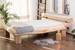 Spokojna sypialnia w stylu Japandi z drewnianym łóżkiem wykonanym z prostych, równych belek, zachowującym czysty i minimalistyczny wygląd. Projekt łóżka eksponuje naturalny rysunek drewna i jest uzupełniony miękką, neutralną pościelą lnianą. Drewniany pieniek pełni funkcję stolika nocnego. W pokoju znajdują się jasne drewniane podłogi, ściany w odcieniach złamanej bieli, delikatne zasłony i minimalistyczne dekoracje, takie jak ręcznie wykonane ceramiczne wazony, jutowy dywan i małe doniczkowe rośliny. Miękkie, naturalne światło wpada przez duże okna, tworząc spokojną i harmonijną atmosferę, która łączy japońską prostotę ze skandynawską przytulnością.
