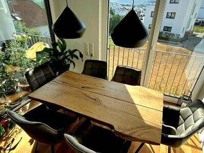 stół z naturalnego drewna, debowy, w salonie połaczonym z jadalnią, urzadzaonym w stylu nowowczesnym, kwiaty w salonie, czarne lampy nad stołem, duze okna z widokiem na podwórko, czarne nowoczesne krzesła z ekoskóry, podłoga dab