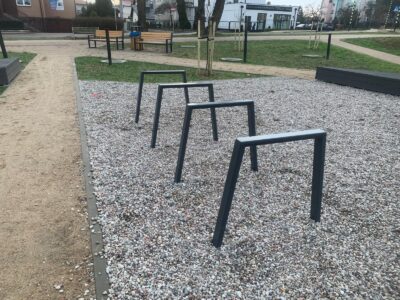 Stalowy stojak rowerowy typu odwrócone U na placu miejskim, zamontowany w podłożu żwirowym wśród małej architektury miejskiej