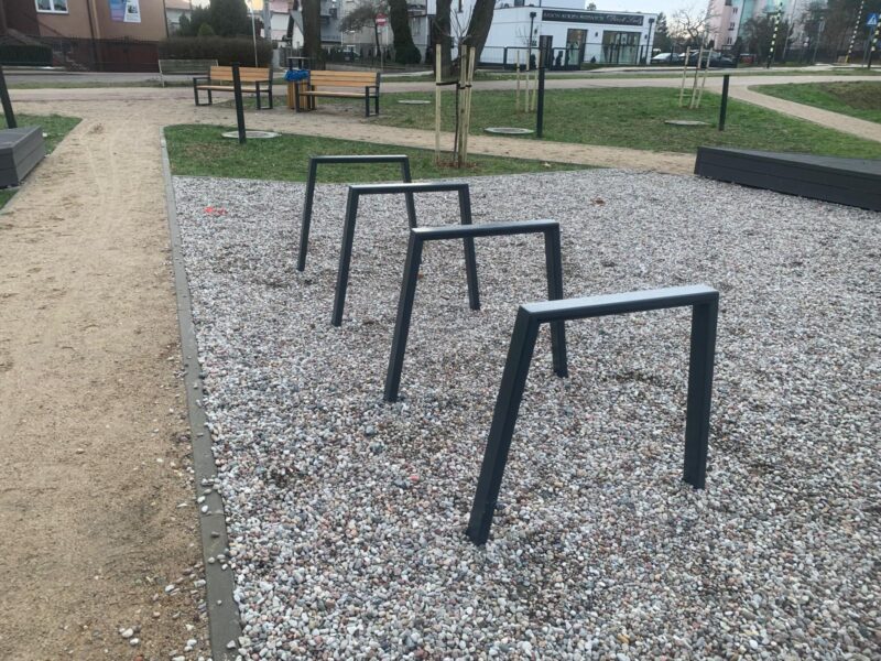 STOJAK NA ROWER (4) Stalowy stojak rowerowy typu odwrócone U na placu miejskim, zamontowany w podłożu żwirowym wśród małej architektury miejskiej