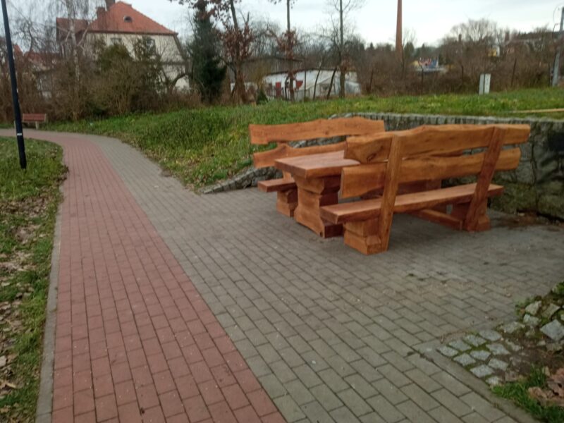lawostol prosty_Kwidzyn Balaton (4) Zestaw ławostołów z naturalnego drewna ustawiony na brukowanej ścieżce przy zbiorniku wodnym. Drewniane ławki i stół o masywnym i rustykalnym charakterze, wkomponowane w otoczenie zieleni i krajobrazu miejskiego z alejkami i budynkami w tle