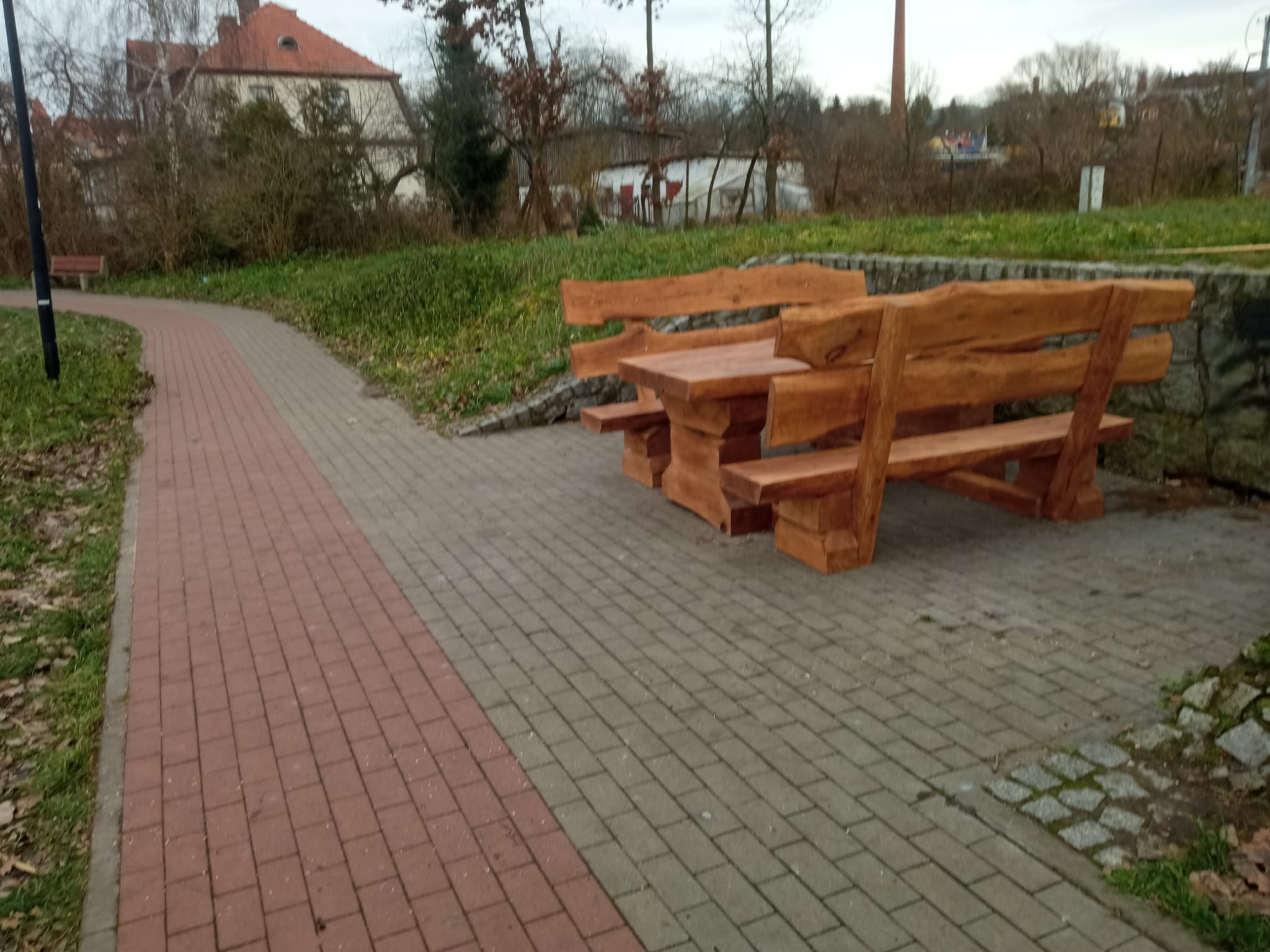 Zestaw ławostołów z naturalnego drewna ustawiony na brukowanej ścieżce przy zbiorniku wodnym. Drewniane ławki i stół o masywnym i rustykalnym charakterze, wkomponowane w otoczenie zieleni i krajobrazu miejskiego z alejkami i budynkami w tle