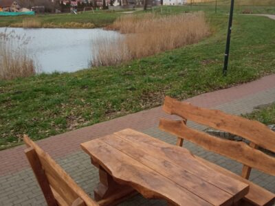 Zestaw ławostołów z naturalnego drewna ustawiony na brukowanej ścieżce przy zbiorniku wodnym. Drewniane ławki i stół o masywnym i rustykalnym charakterze, wkomponowane w otoczenie zieleni i krajobrazu miejskiego z alejkami i budynkami w tle