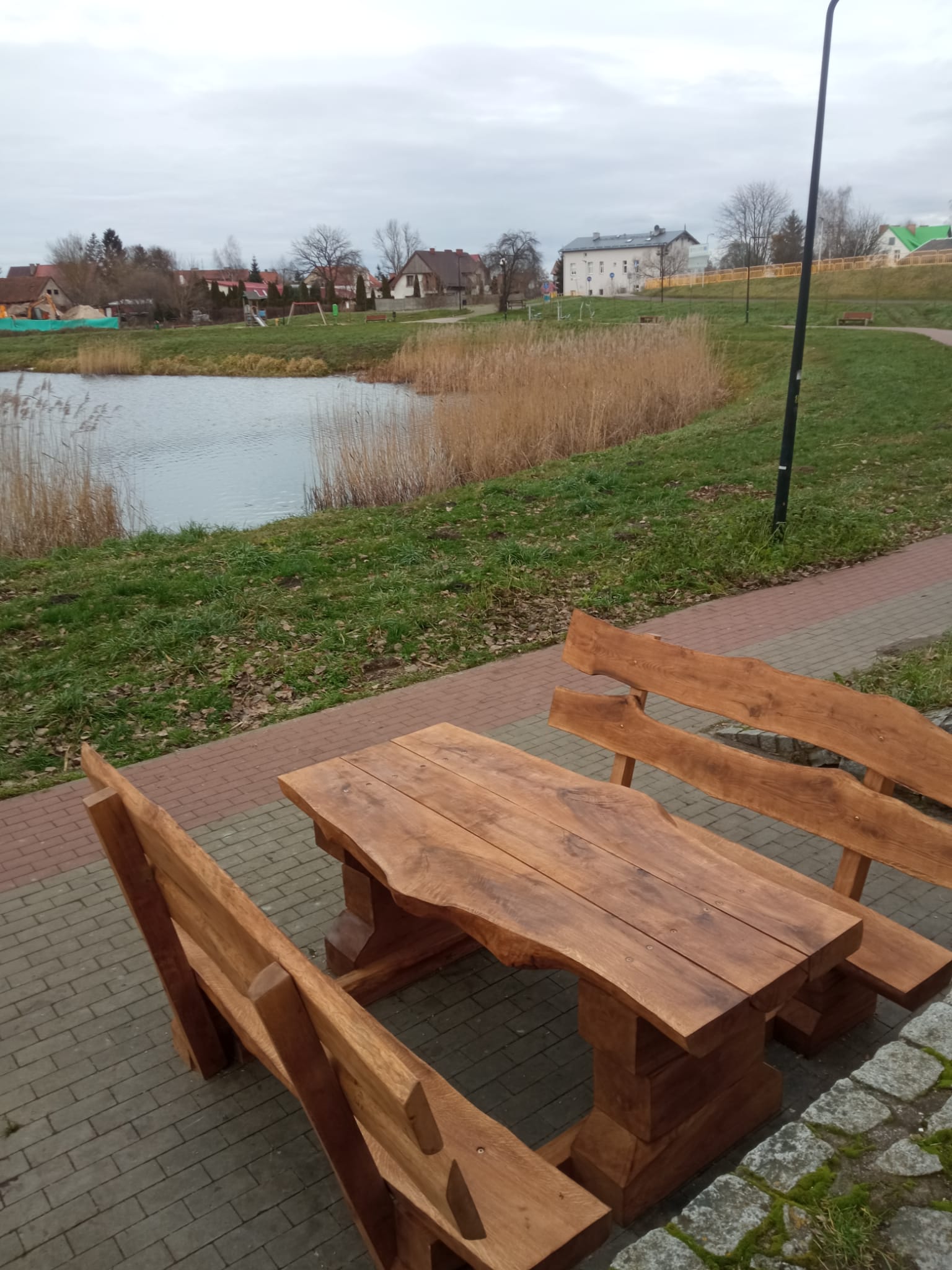 Zestaw ławostołów z naturalnego drewna ustawiony na brukowanej ścieżce przy zbiorniku wodnym. Drewniane ławki i stół o masywnym i rustykalnym charakterze, wkomponowane w otoczenie zieleni i krajobrazu miejskiego z alejkami i budynkami w tle