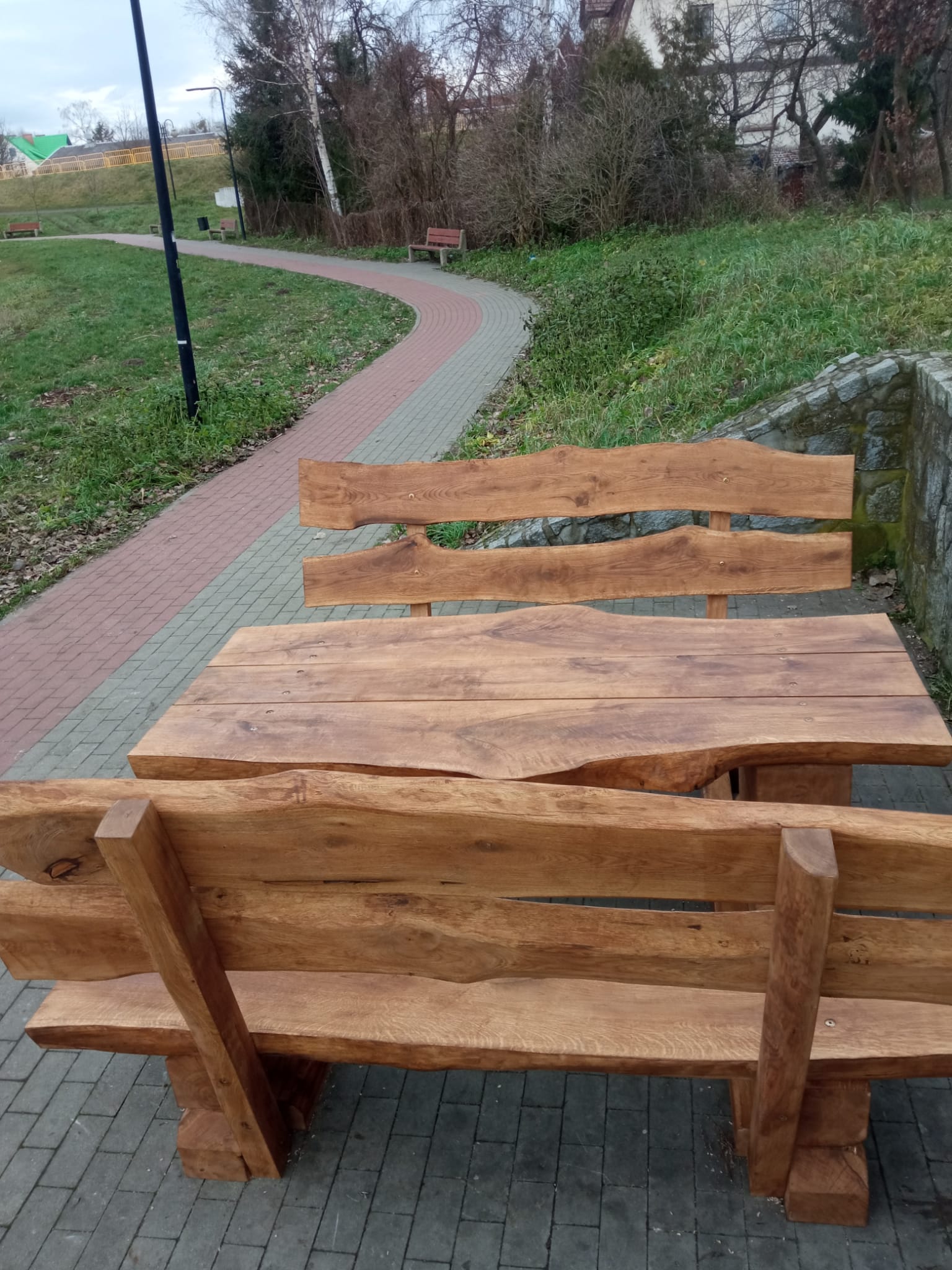 Zestaw ławostołów z naturalnego drewna ustawiony na brukowanej ścieżce przy zbiorniku wodnym. Drewniane ławki i stół o masywnym i rustykalnym charakterze, wkomponowane w otoczenie zieleni i krajobrazu miejskiego z alejkami i budynkami w tle