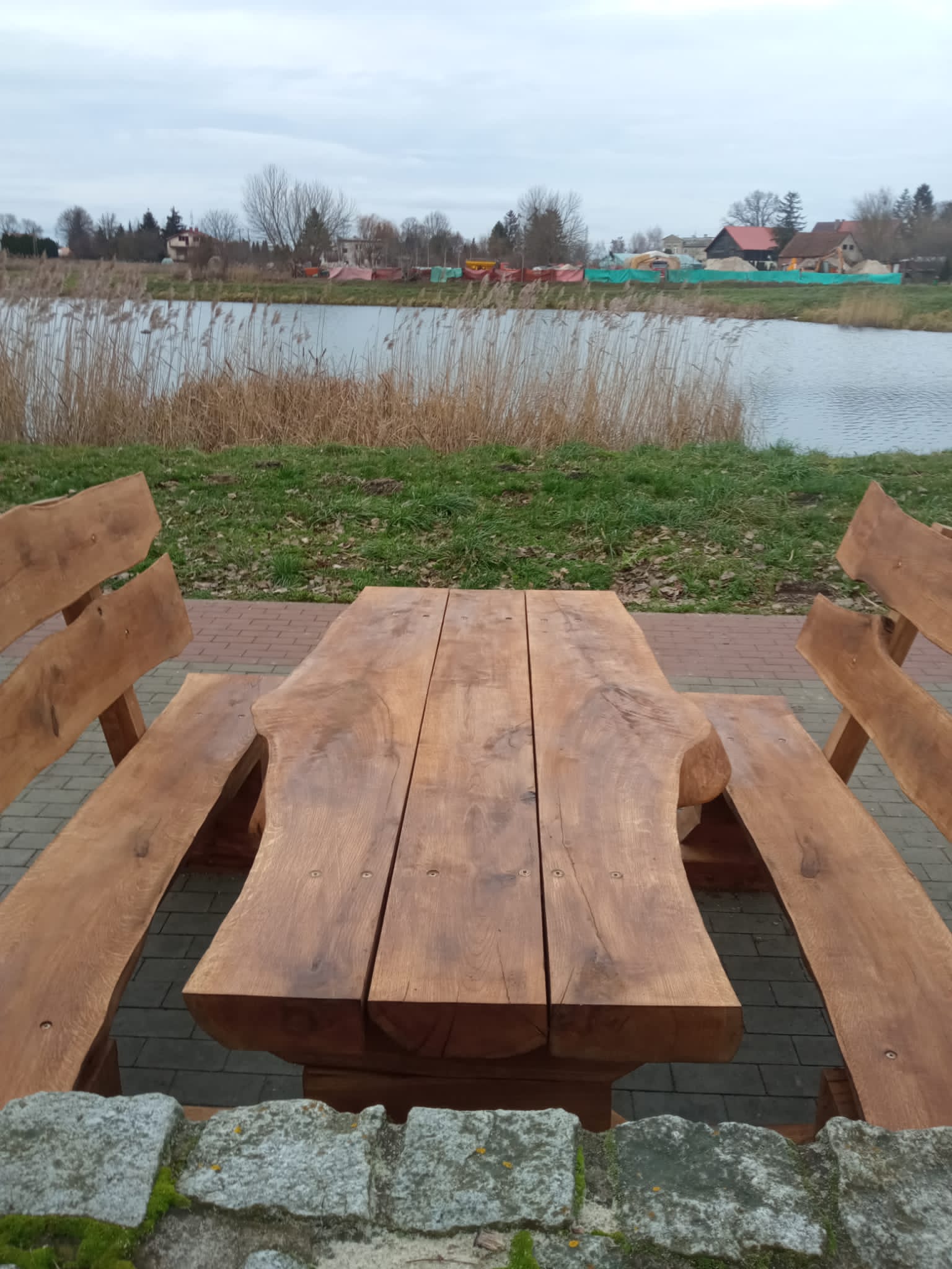 Zestaw ławostołów z naturalnego drewna ustawiony na brukowanej ścieżce przy zbiorniku wodnym. Drewniane ławki i stół o masywnym i rustykalnym charakterze, wkomponowane w otoczenie zieleni i krajobrazu miejskiego z alejkami i budynkami w tle