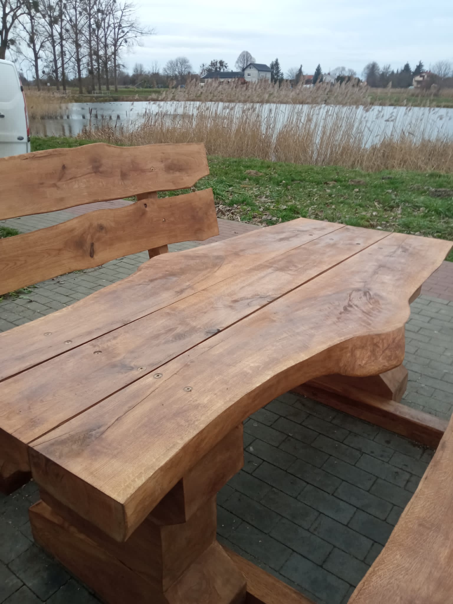 Zestaw ławostołów z naturalnego drewna ustawiony na brukowanej ścieżce przy zbiorniku wodnym. Drewniane ławki i stół o masywnym i rustykalnym charakterze, wkomponowane w otoczenie zieleni i krajobrazu miejskiego z alejkami i budynkami w tle