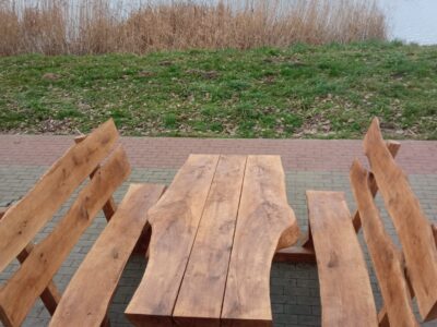 Zestaw ławostołów z naturalnego drewna ustawiony na brukowanej ścieżce przy zbiorniku wodnym. Drewniane ławki i stół o masywnym i rustykalnym charakterze, wkomponowane w otoczenie zieleni i krajobrazu miejskiego z alejkami i budynkami w tle