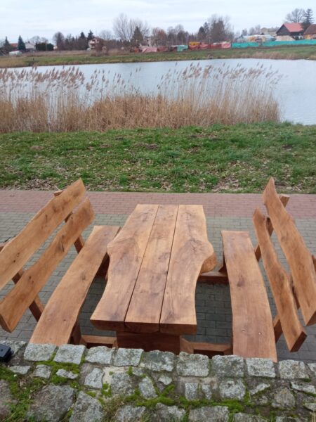 Zestaw ławostołów z naturalnego drewna ustawiony na brukowanej ścieżce przy zbiorniku wodnym. Drewniane ławki i stół o masywnym i rustykalnym charakterze, wkomponowane w otoczenie zieleni i krajobrazu miejskiego z alejkami i budynkami w tle